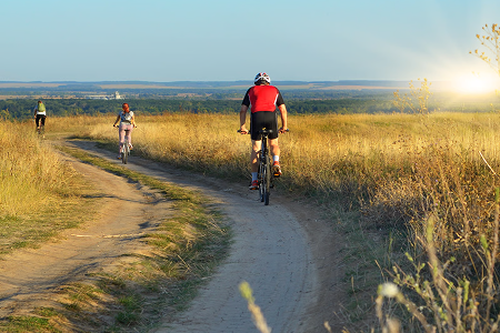 Cyclists ride along a dirt path through a sunlit grassy field, with a bright horizon in the background, evoking a sense of freedom and adventure.