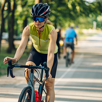 A woman in a yellow cycling jersey rides a bicycle on a tree-lined path. She wears a helmet and sunglasses. A cyclist in blue follows behind.