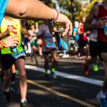 A runner's hand reaches out towards a group of marathoners, offering a drink in a blue and black cup. The scene is vibrant and energetic, with runners blurred in motion.